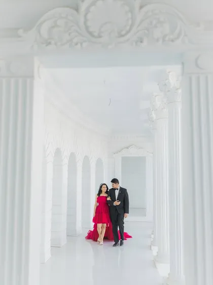 An expecting couple shares a quiet moment while walking through a majestic white corridor. This wide shot captures the grandeur of the architecture, framing them and her striking red dress in a truly cinematic style.