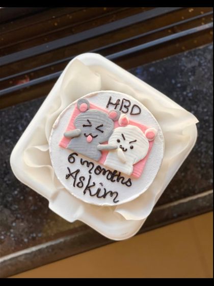 An adorable cartoon-themed bento cake celebrating a 6-month anniversary. It features two cute characters with the message "HBD 6 months Askim."