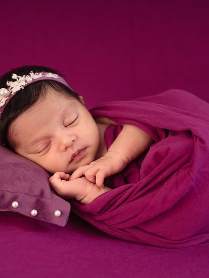 A close-up of a peacefully sleeping baby in a rich purple wrap, highlighting the delicate pearl headband and the serene expression.