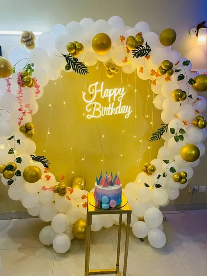 A simple and elegant birthday photo corner with a circular balloon arch in white and gold, a neon sign, and a plinth for the cake.