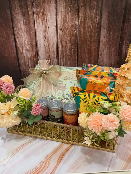 This Rakhi snack hamper combines delicious treats with elegant packaging, including a beautiful box and floral arrangements in a golden basket.