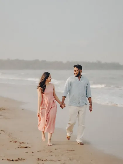 A candid shot of Anushka and Manu sharing a look while walking along the shoreline. The warm, earthy tones of the sand and soft light complement their relaxed outfits.