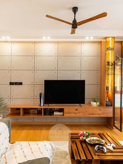 This living room features a unique TV unit with an integrated pooja nook. The concrete-finish wall panels contrast beautifully with the warm wood of the console and the backlit glass pooja cabinet.