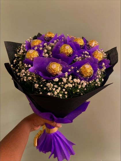 The purple Ferrero Rocher bouquet held up to display its full form, showing the contrast between the vibrant purple, the dark wrapping, and the delicate white flowers.
