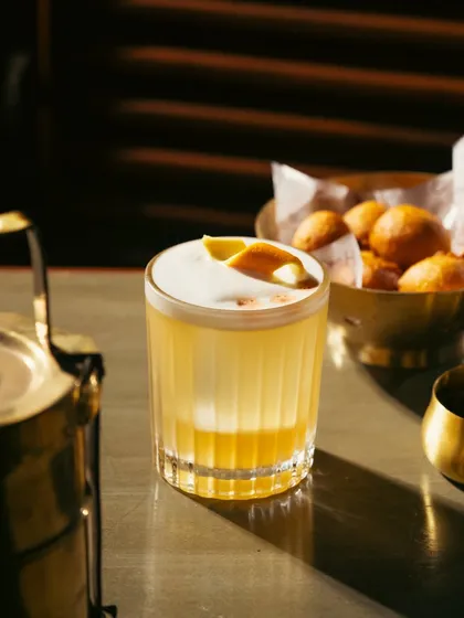 A classic whiskey sour with a foam top and orange peel garnish. The lighting highlights the texture of the foam and the rich color of the drink.
