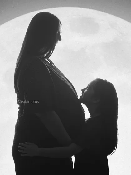 A powerful and creative black and white silhouette. This image tells a story of a growing family, with an older sibling looking up at her mother's baby bump against a full moon backdrop, symbolizing the new light coming into their lives.
