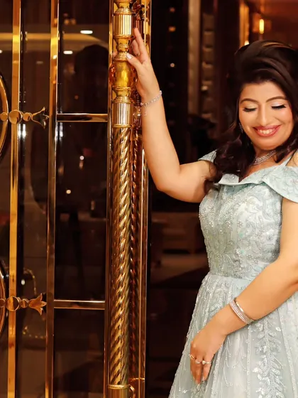 A smiling woman in a beautiful light blue gown poses against an ornate golden door, a perfect shot from an elegant anniversary or engagement party.