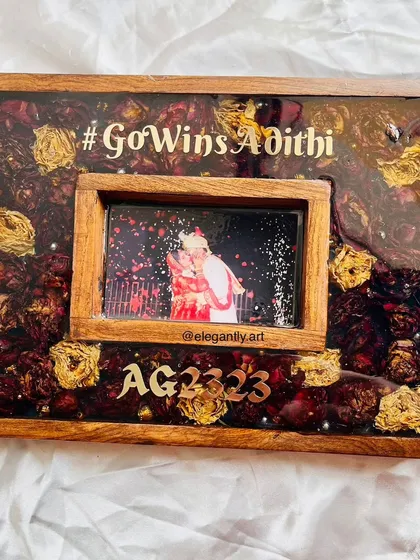 This rectangular teakwood frame features a unique floating photo design, surrounded by preserved red and gold roses from a wedding garland. The couple's hashtag and initials add a modern, personalized touch.