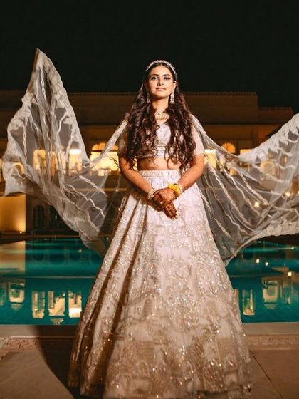 The bride stands powerfully by the pool, her cape-like dupatta flowing in the wind, creating a majestic Sangeet look.
