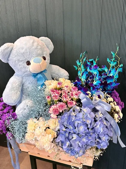 A different angle of the blue-themed teddy bear and flower crate. The mix of textures between the soft bear, delicate petals, and rustic wooden crate makes it a visually interesting gift.