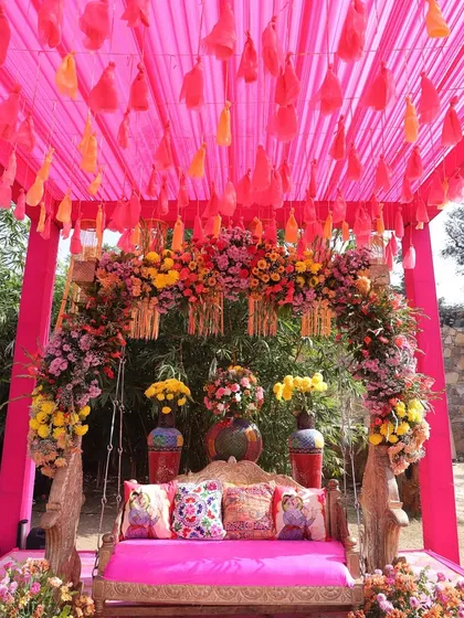A vibrant pink jhoola (swing) seating for a Mehendi, set under a canopy of pink drapes and surrounded by a riot of colorful flowers.