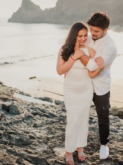 A wide shot of the couple embracing on a rocky outcrop with the beach and ocean behind them. This photo combines their intimate moment with the grandeur of the coastal landscape.