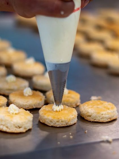 A close-up of our pastry team at work. A chef pipes delicate swirls of cream onto freshly baked scones, a small but crucial detail for an unforgettable high tea experience.
