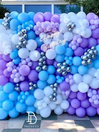 An outdoor balloon wall creating a vibrant photo backdrop. The mix of blue, purple, white, and silver balloons provides a fresh and modern look for any celebration.
