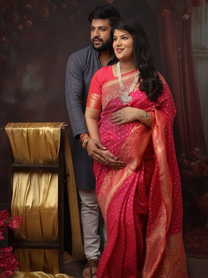 A beautiful couple portrait celebrating motherhood with elegance. The mom-to-be glows in a bright pink saree, her partner standing lovingly behind her.