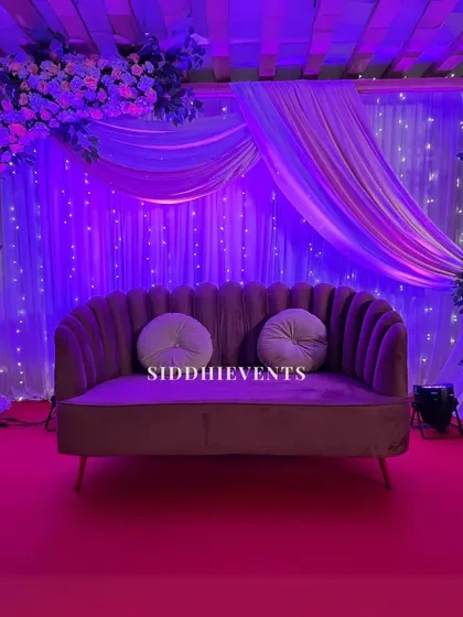 A dramatic and moody stage setup with deep purple lighting, sheer drapes with fairy lights, and floral arrangements, creating an enchanting atmosphere for an evening event.