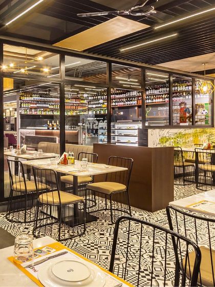This section of the restaurant features striking black and white patterned floor tiles that contrast with the warm wood tones and simple metal chairs. A glass partition provides a view of the well-stocked bar area, adding to the open feel.