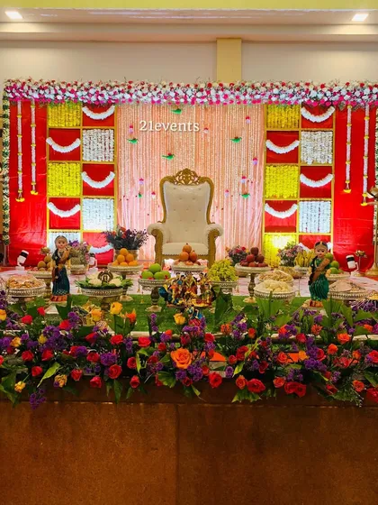 A classic Seemantha decor setup with a red, yellow, and white color scheme. The stage includes a central throne, traditional dolls, and a flowerbed, making it a perfect setting for the ceremony.