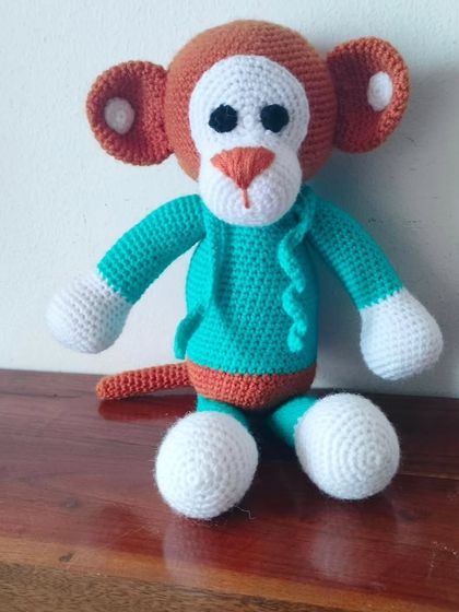 A handmade crochet monkey in a sea green shirt. He has a long tail and a very friendly face.
