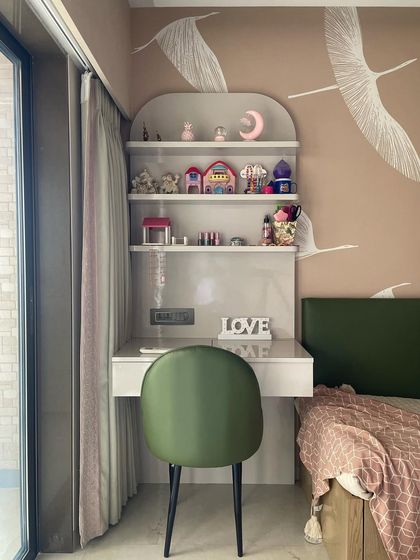 A charming study and vanity area in a girl's room. The design features a built-in desk with arched shelving, a soft green chair, and a whimsical bird-themed wallpaper.