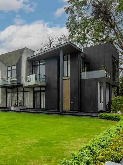 An angled view of a modern farmhouse, showcasing its geometric massing against a backdrop of mature trees. The design uses contrasting textures and volumes to create a powerful architectural statement in a natural setting.