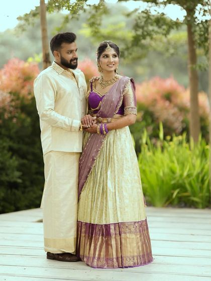 A couple posing outdoors in their engagement attire. Her off-white and purple half saree is complemented by his cream silk dhoti and kurta, creating a harmonious look.