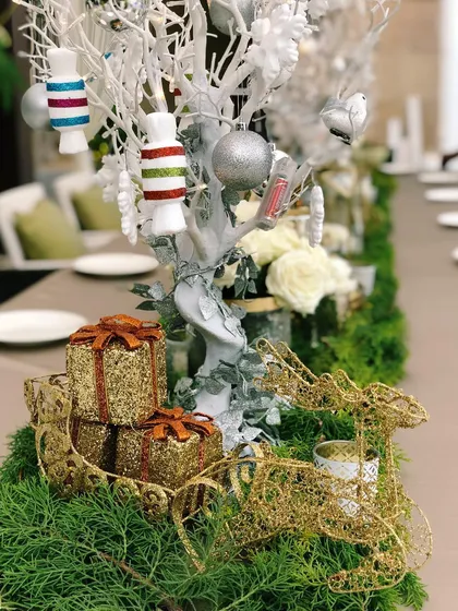 A close-up of a Christmas Eve dinner tablescape I designed. The centerpiece includes a white decorative tree with candy ornaments, surrounded by a lush green runner, glittery gold gift boxes, and a delicate wire reindeer, creating a festive and elegant holiday setting.