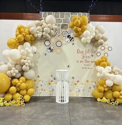 A sweet "Our little bee is on the way" baby shower backdrop, featuring a honeycomb pattern and a beautiful yellow, white, and beige balloon arch.
