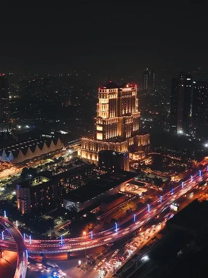 A long exposure aerial photograph of Kolkata at night, capturing the light trails from traffic flowing around the ITC Royal Bengal. This technique transforms a busy city scene into a dynamic and artistic image, showcasing the urban energy from a unique perspective.