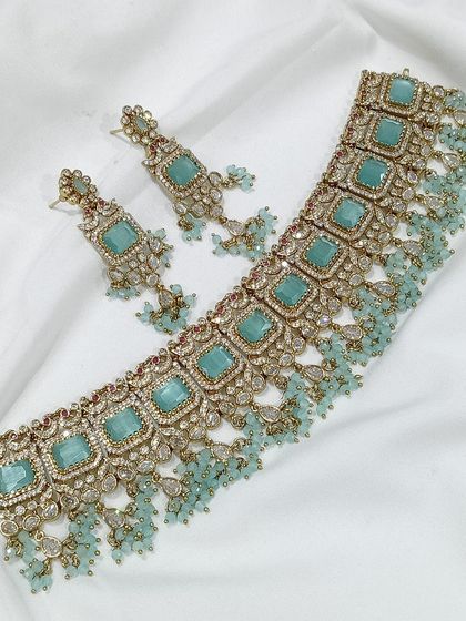 A popular choker design featuring square-cut stones, available in a variety of colors like emerald green, white, aqua blue, and baby pink. The matching earrings complete this elegant and modern ethnic set.