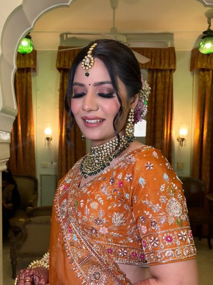A smiling bride, ready for her wedding. Her hair is styled in a beautiful floral bun, with soft tendrils framing her face, complementing her glowing makeup and heavy jewelry.