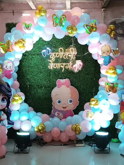 This baby shower photo booth features a circular balloon arch against a green wall backdrop. The theme combines cute baby cutouts with traditional Radha Krishna figures.