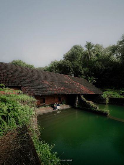 A wide shot showing the couple in the serene, rustic setting of a traditional Kerala house by the water. This pre-wedding shoot highlights the beauty of heritage architecture.
