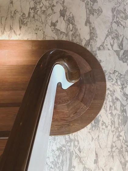 A top-down view of the whirling staircase against a backdrop of Carrara marble. This detail highlights the contrast between the organic curve of the wooden handrail and the natural pattern of the stone.
