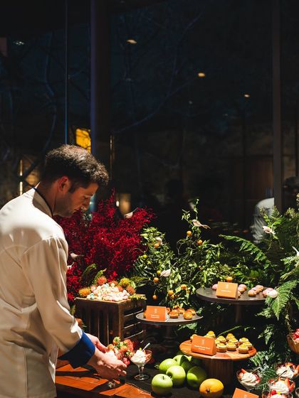 Chef Maxime Montay adds the final touches to an exquisite grazing table. The display is a forest of flavors, with fresh fruits, miniature desserts, and vibrant floral arrangements.