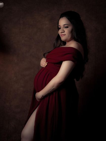 A dramatic solo portrait featuring a deep red, off-the-shoulder gown. The lighting is carefully controlled to create a moody, intimate atmosphere that emphasizes the mother's form.