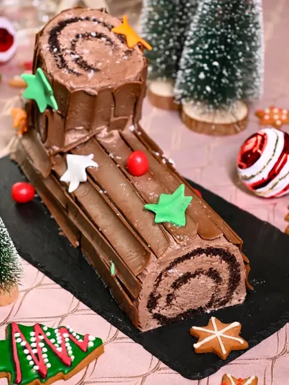 A classic Christmas Yule log cake, decorated with festive sugar stars and set among gingerbread cookies and mini trees. This image evokes a cozy, traditional holiday feeling.