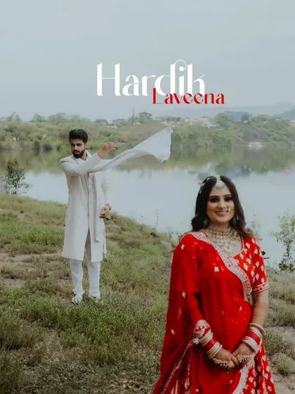 A beautifully composed pre-wedding portrait by a lake, with the bride in a vibrant red lehenga and the groom in the background.