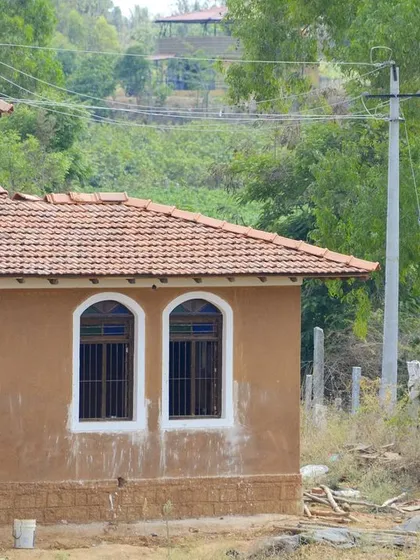 The mud house during the final stages of construction, with workers applying the finishing touches. The home sits gently on the land, surrounded by the natural landscape of the farm.