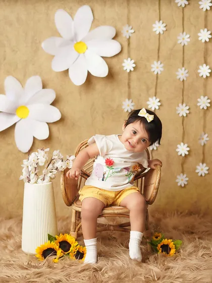A cheerful baby girl in a daisy-themed setup. Her bright smile and the sunny yellow shorts create a happy and vibrant milestone photo.