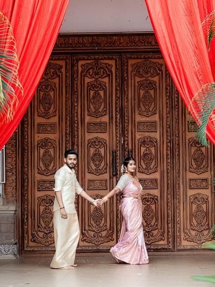 A grand entrance shot of a couple holding hands, framed by beautiful red curtains and lush greenery. We make your wedding portraits look majestic.