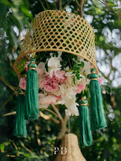 A beautiful decor detail for a boho Mehendi: a rattan basket filled with flowers and adorned with green tassels, hanging from a tree.