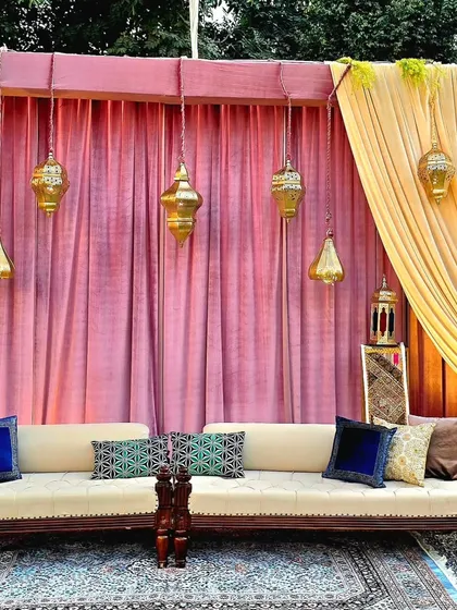 This seating arrangement for a Sangeet night features a long sofa adorned with a variety of patterned and solid-colored cushions. The backdrop of pink drapes and hanging Moroccan lanterns creates a rich, festive atmosphere.