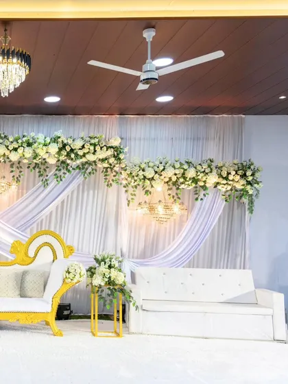 A classic and elegant stage setup in white and gold. The backdrop features white drapes and floral arrangements, complemented by a mix of modern and traditional seating.