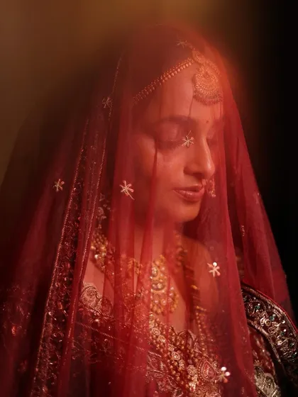 A dreamy, artistic shot of Laxmi through her red veil. This image perfectly captures the soft, ethereal quality of her traditional bridal makeup, highlighting a timeless and graceful aesthetic.