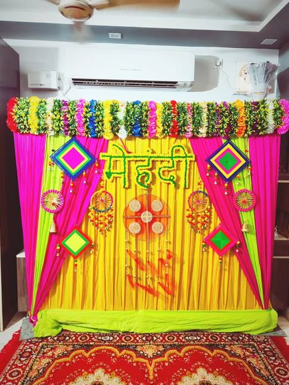 A vibrant indoor Mehendi backdrop with pink, yellow, and green drapes, a floral valance, and traditional hangings.