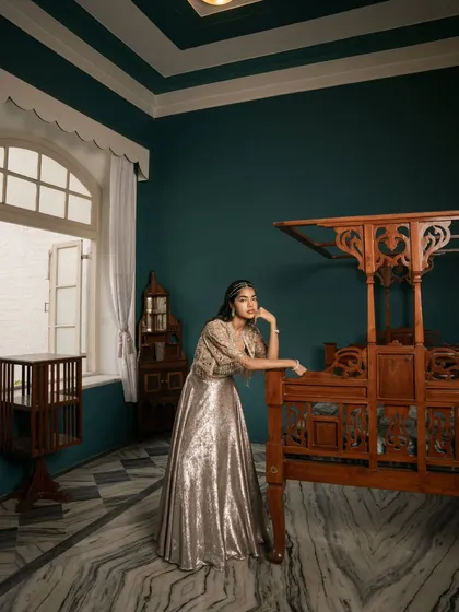 This environmental portrait places the model and her shimmering lehenga in a beautifully appointed room, telling a story of heritage and modern luxury. The four-poster bed adds a classic, regal element.