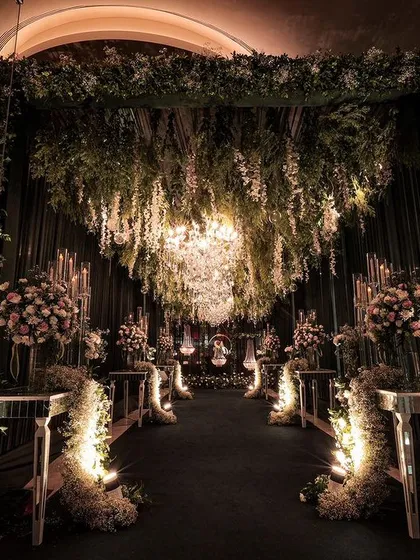 An absolutely magical entrance tunnel for a reception at Pullman New Delhi. The design features a ceiling of hanging florals and greenery, with a large chandelier and mirrored pedestals creating a forest of light.