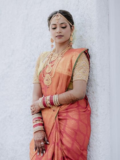 A stunning portrait of the bride leaning against a white wall. The simplicity of the background makes her vibrant red and orange saree stand out.