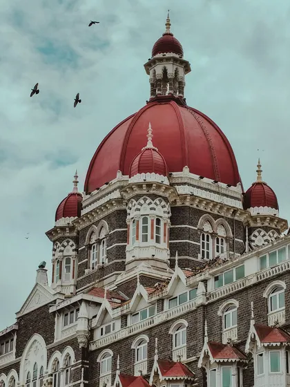 The famous red dome of the Taj Mahal Palace hotel, with birds flying past against a cloudy sky. A truly iconic Mumbai frame.